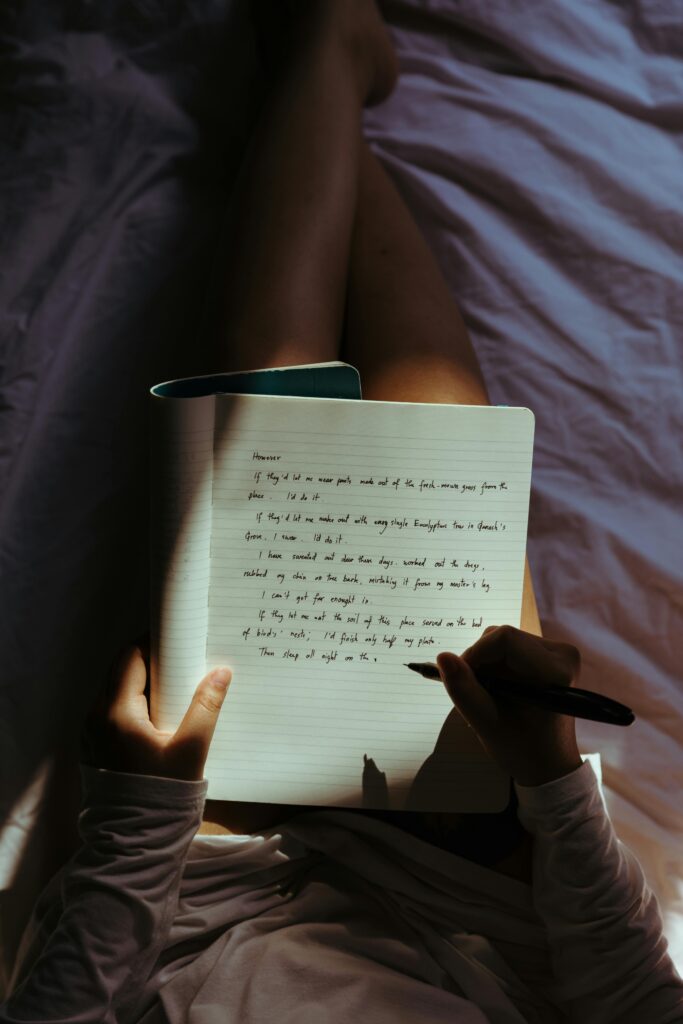 pexels-photo-4132327-4132327 Woman writing in a notebook on her lap while relaxing comfortably in a cozy, dimly lit bedroom setting.
